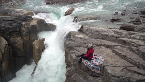 female sitting on rock above waterfall and glacial river in canadian countryside on cold autumn day