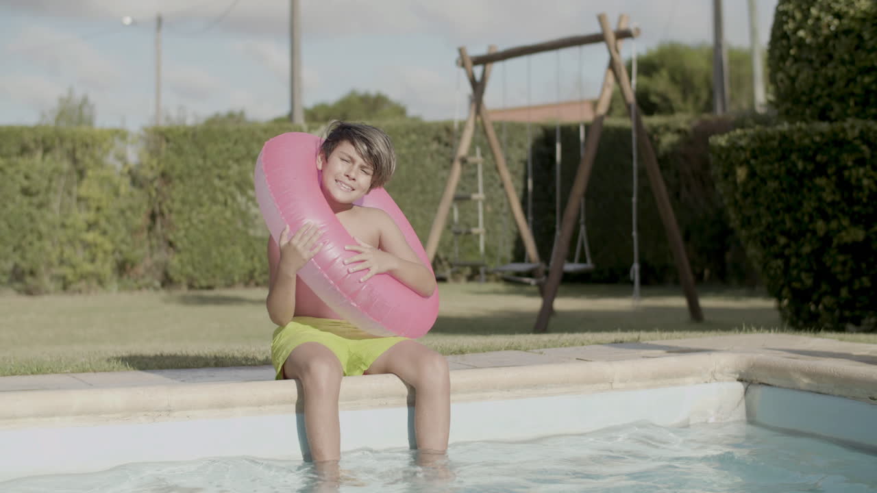 una foto larga de un niño con un anillo inflable rosa sentado a la orilla de la piscina.
