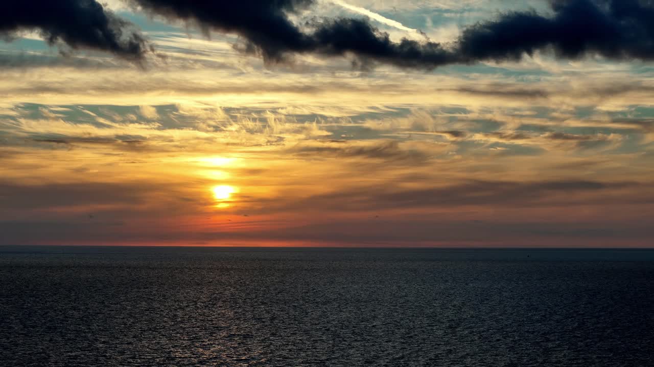 Sonnenlicht, das durch dunkle Regenwolken über dem mediterranen Horizont bricht, während eines Wintersonnenuntergangs auf Gozo, Malta, mit goldenen Farbtönen, die sich auf dem ruhigen, tiefblauen Meer spiegeln.