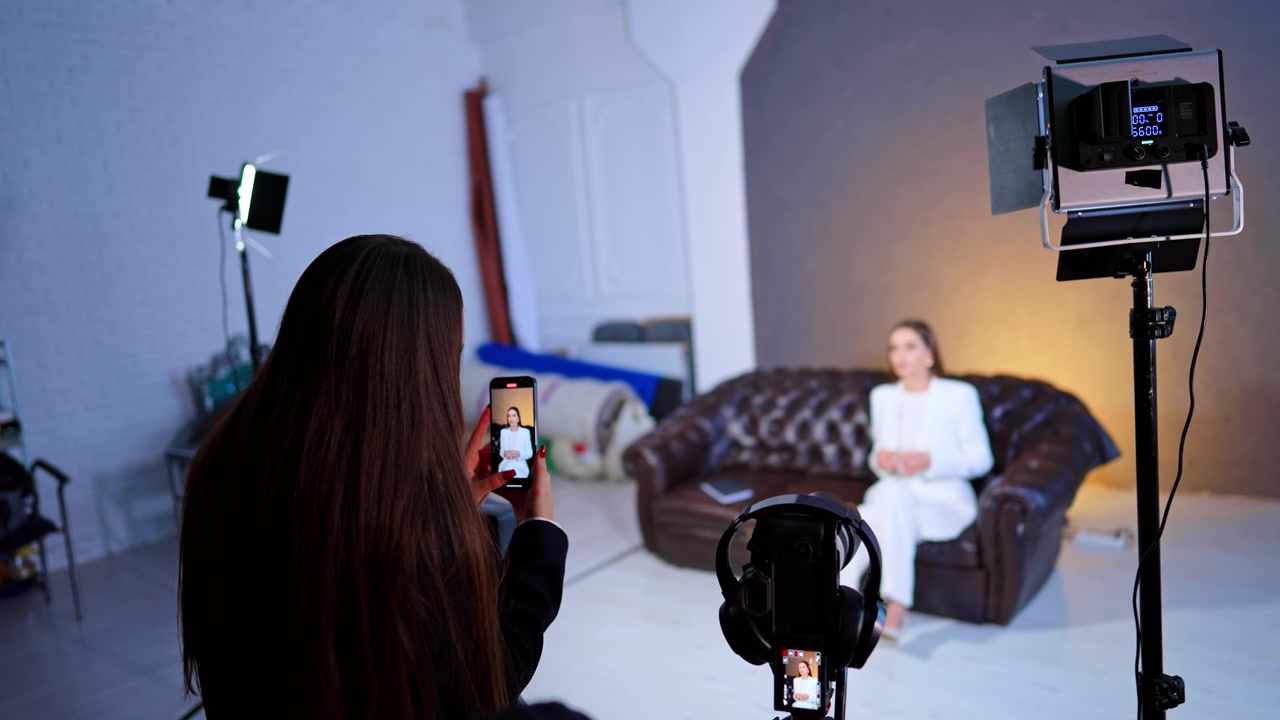 Asistan Stüdyoda Akıllı Telefonla Röportaj Konusunu Kaydediyor. Stüdyoda Beyaz Takım Elbiseli Oturan Kadını Kamera ve LED Işıkla Akıllı Telefona Kaydeden Asistan.