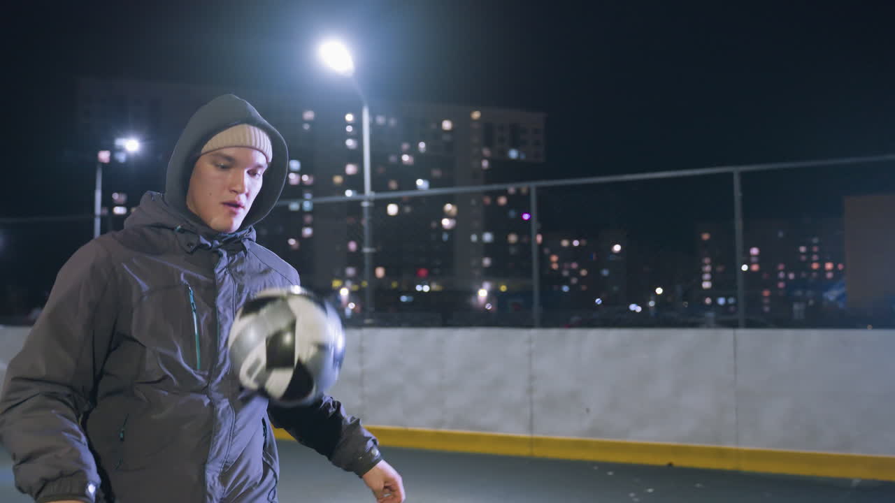 Athlete joggling football with his knee during nighttime training session on urban field, surrounded by illuminated streetlights and residential buildings in the background