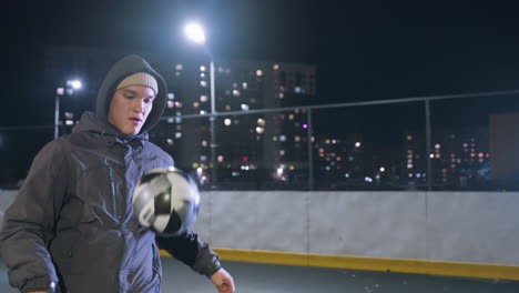 atleta jugando al fútbol con su rodilla durante una sesión de entrenamiento nocturna en un campo urbano, rodeado de farolas iluminadas y edificios residenciales en el fondo