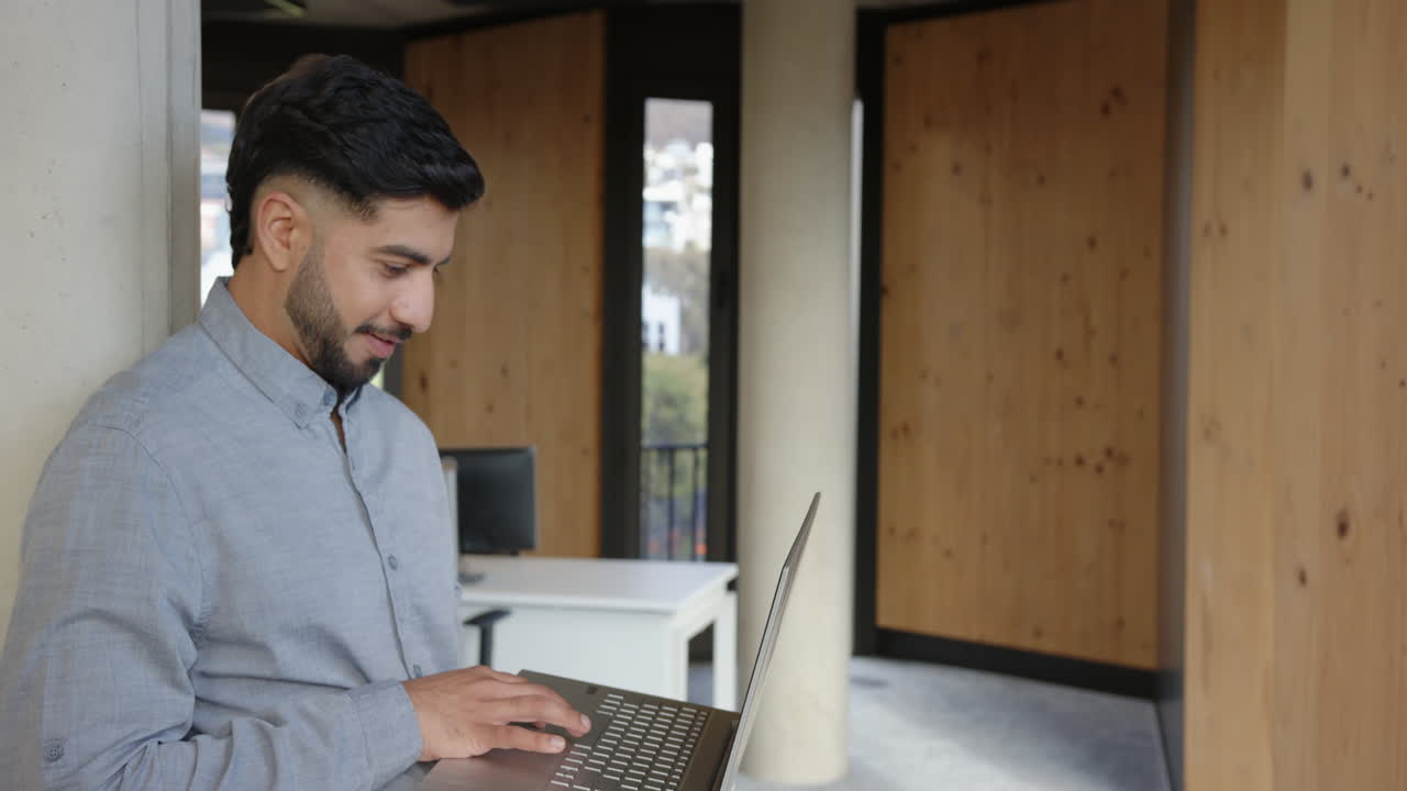 Using laptop, Indian man standing in modern office, concentrating on work