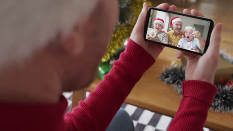 hombre caucásico con sombrero de santa usando un teléfono inteligente para una videollamada de navidad con una familia sonriente en la pantalla