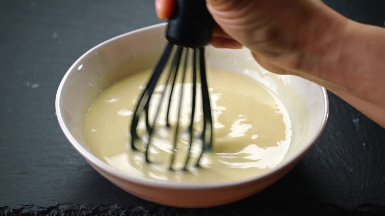 Whisking hand stirring batter in bowl with whisk entering from top right on counter smoothing