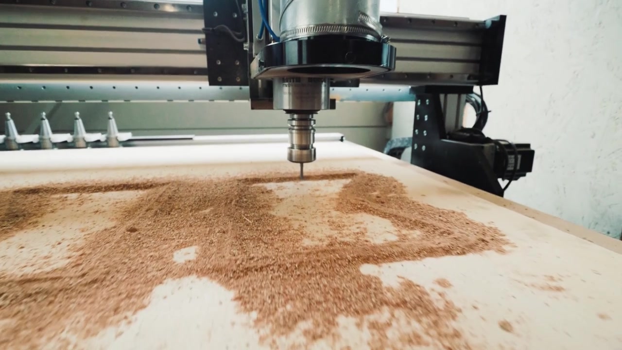 Ampia ripresa dal basso di una macchina CNC che taglia una grande tavola di compensato coperta di segatura all'interno di una falegnameria