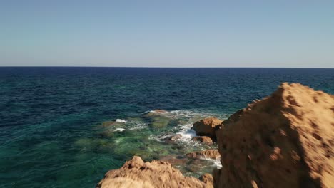 Toma-Aérea-De-Una-Costa-Con-Hermosas-Aguas-Turquesas-Del-Mar-Mediterráneo-Salpicando-En-Las-Rocas-De-Color-Rojo