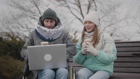 disabled man in wheelchair and his friend watching something funny on laptop computer while drinking takeaway coffee at urban park in winter 3