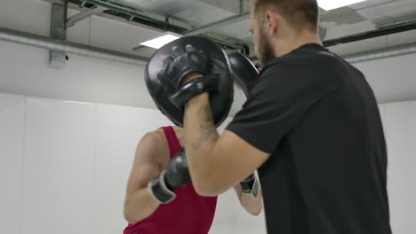 at a boxing training session in the white room a man beats his gloved hands on the coach's paws. perform deviations and dives from the blows.