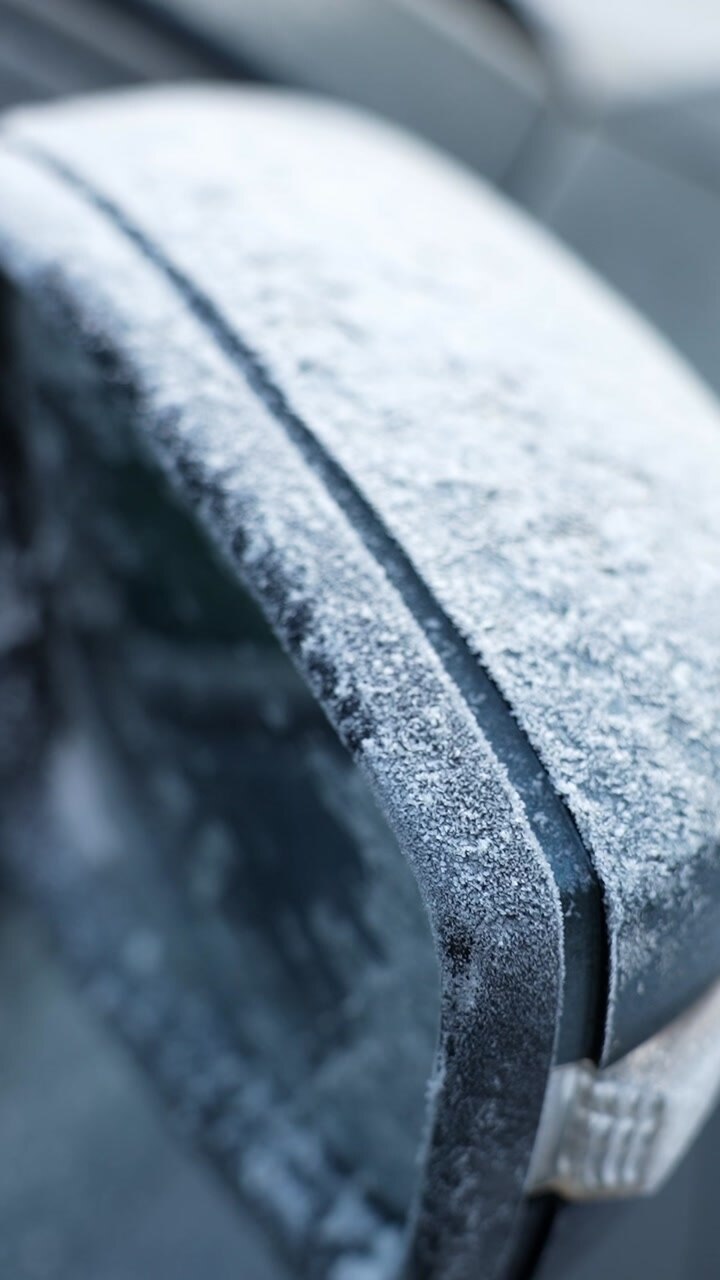 Vertical macro close-up of a car side mirror coated with frost crystals on a cold winter morning, shallow depth of field and soft bokeh for weather, safety, and automotive themes.