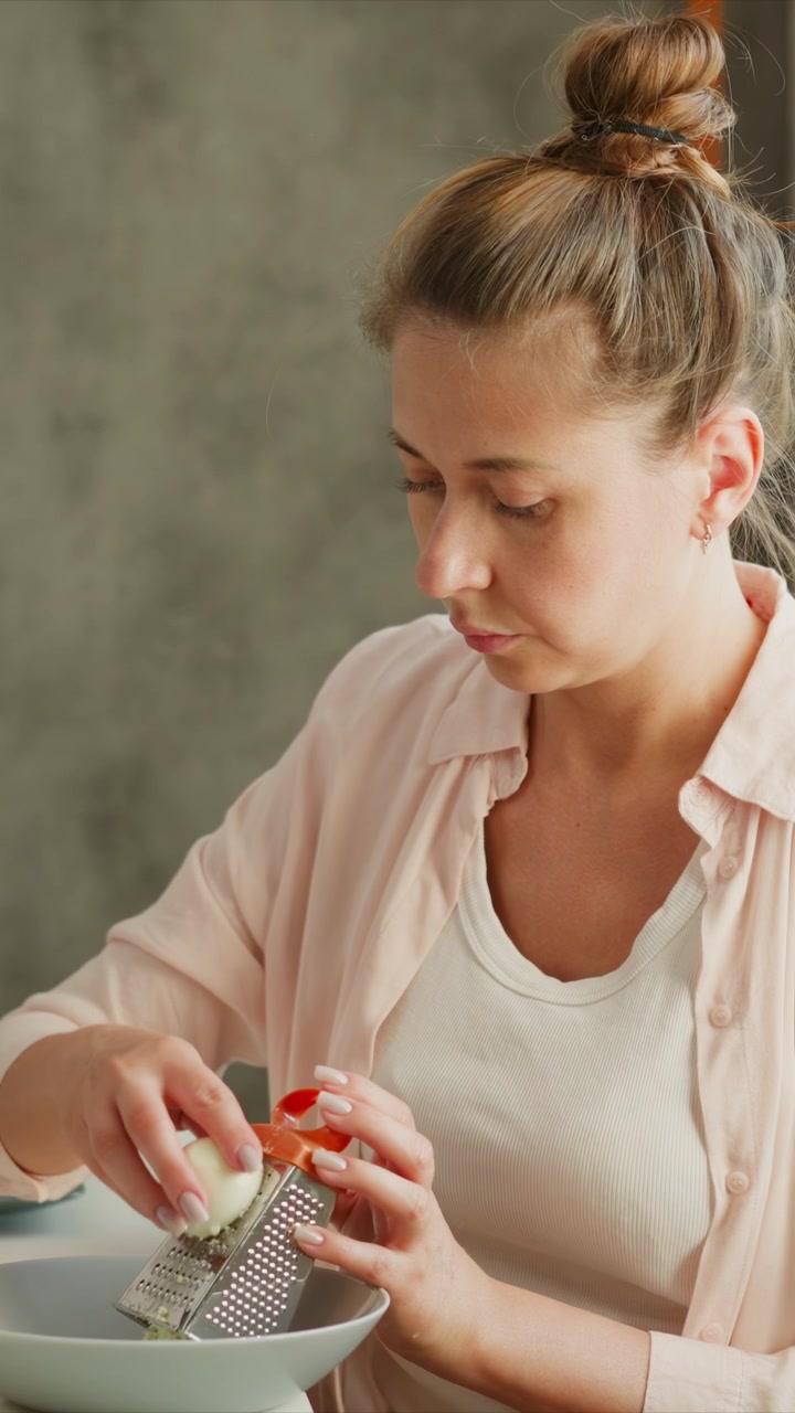 Una donna grattugia una cipolla in una ciotola in una luce naturale soffusa. La donna indossa una camicia rosa sopra un top bianco, con i capelli raccolti e smalto argentato visibile mentre tiene ferma una grattugia di metallo.