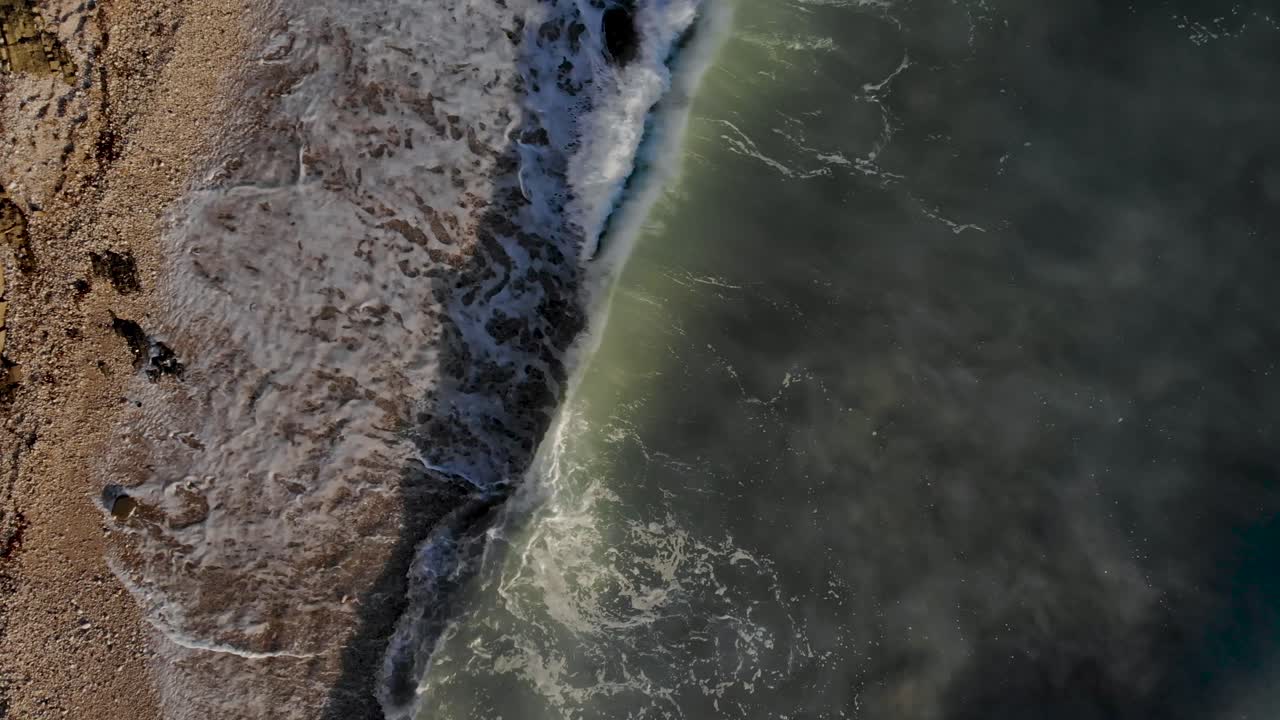 морские волны пенятся и плещутся на галечном пляже, спускающемся со скалистого холма в средиземноморье