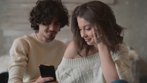 Linda-Pareja-Joven-Sentada-En-La-Cama-Mirando-Un-Teléfono-Y-Usando-Ropa-De-Invierno