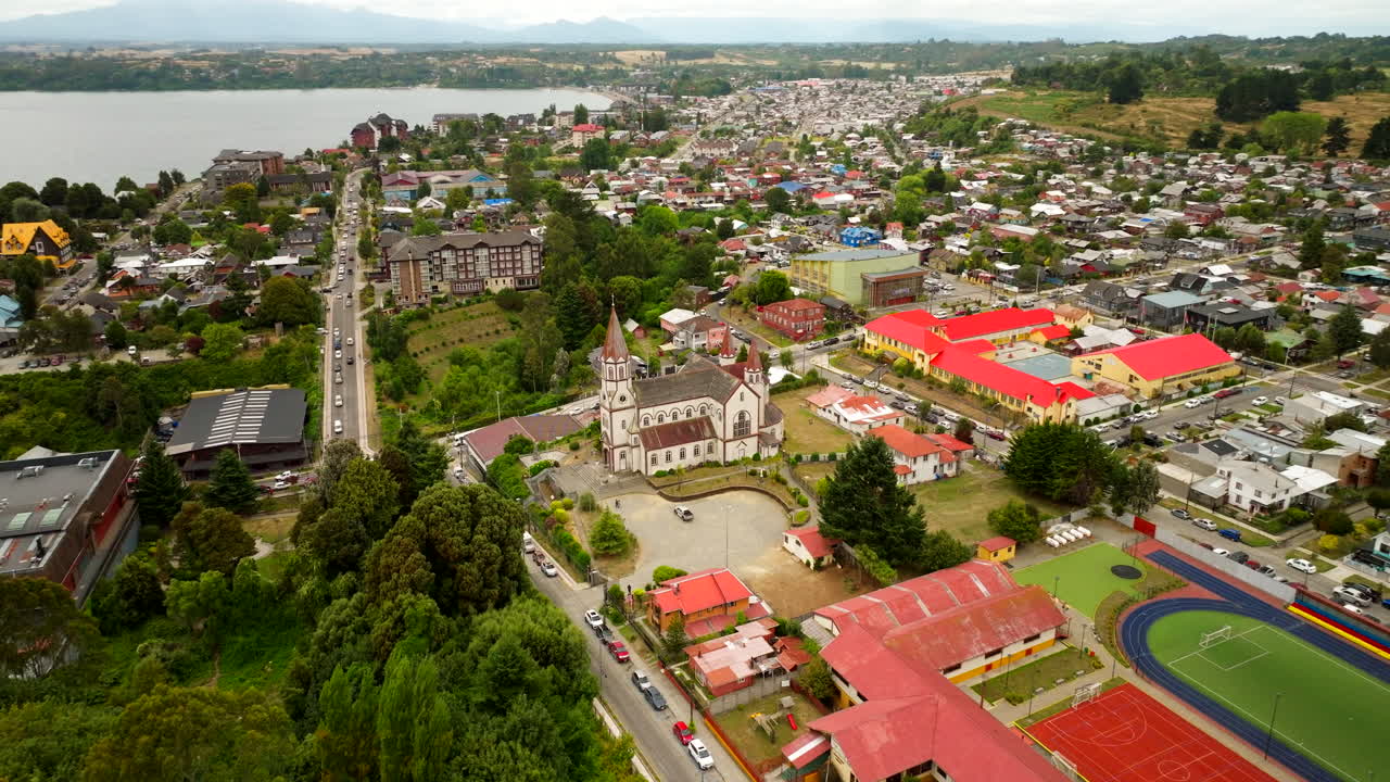 Aerial orbital view of Puerto Varas, Chile, featuring a red-roofed church, sports field, and colorful buildings surrounded by trees and mountains on a cloudy summer day