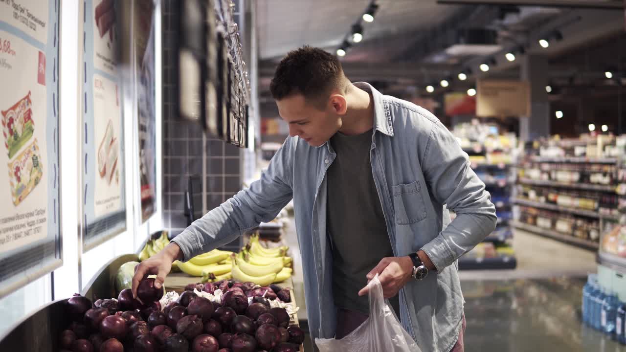 imágenes laterales de un hombre caucásico elige cebollas rojas en un supermercado. el tipo alto positivo con camisa azul compra alimentos y verduras en la tienda de comestibles. cámara lenta