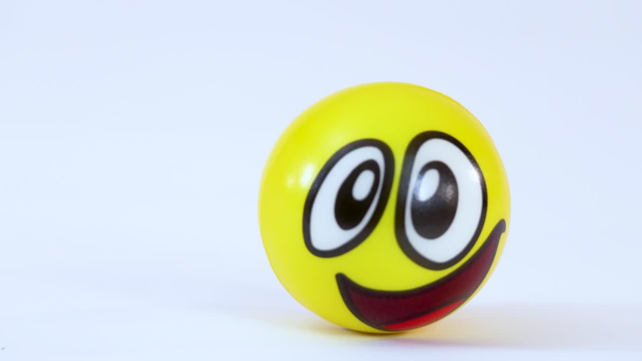 Una palla antistress emoji gialla rimbalza nell'inquadratura e rotola su uno sfondo bianco da studio.