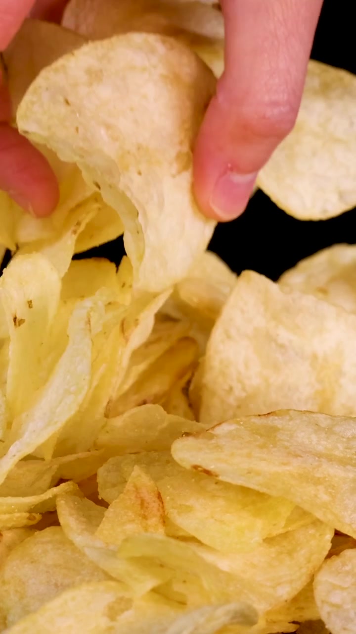 A hand reaches into a pile of golden potato chips against a dark background.
