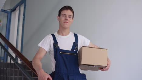 a courier in a white t-shirt and blue overalls stands on the stairs in a high-rise building holding a box in one hand