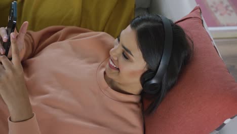 video of happy biracial woman lying on sofa with headphones and using smartphone