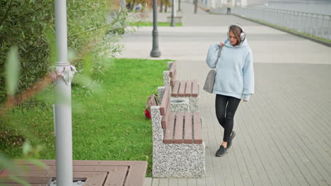 female adult walking majestically to relax on concrete bench, holding handbag, with blurred man walking beside iron railing in background, leaves, poles, serene park atmosphere