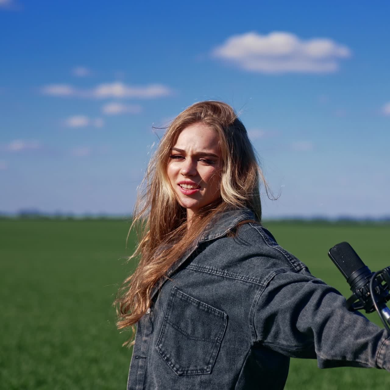 Beautiful girl singer in a green field. Attractive blond woman singing into a microphone and energizing looking at the camera