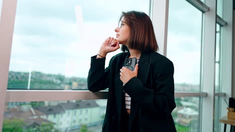 thoughtful caucasian brunette stands looking at window. business lady holding a phone and waiting for the call. low angle view.