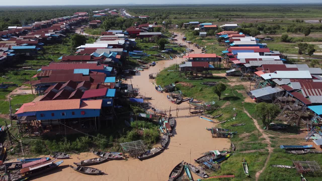Kamboçya kırsalında, tekneler, ahşap yürüme yolları ve kırsal yerleşim manzarası ile çamurlu nehir kanalı boyunca sıralanan geleneksel kazık evleri gösteren havadan drone görüntüsü