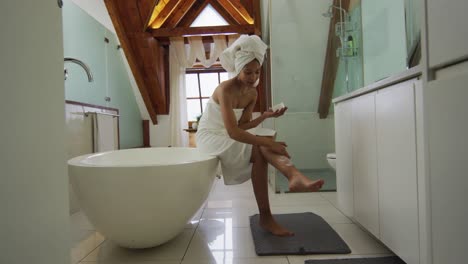 mixed race woman wearing towel on head applying cream on her legs