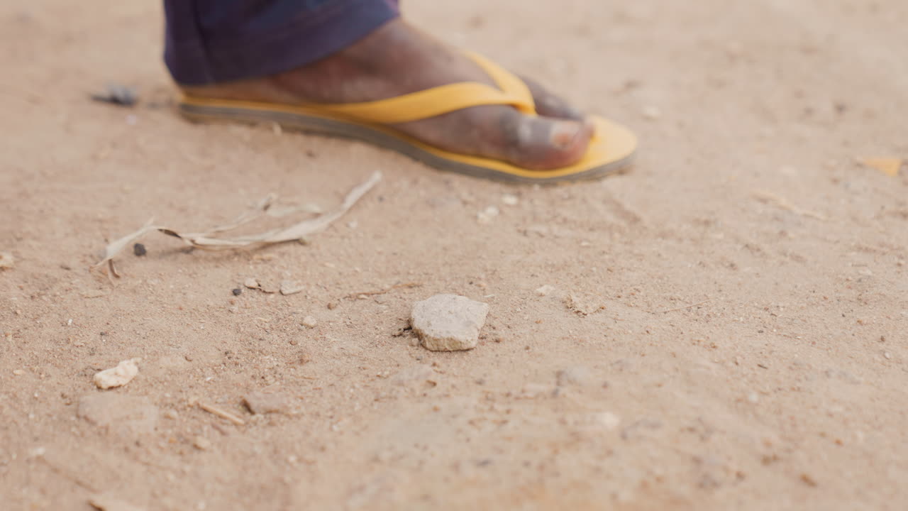 Primo piano di un sandalo giallo su terreno polveroso che mostra un piede usurato che calpesta ciottoli e terra asciutta, un passo lento e deliberato, calda luce solare, terra dalla superficie ruvida, scena di strada quotidiana, umile resilienza.