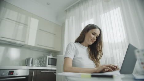 mujer feliz trabajando en una computadora portátil en la cocina. persona femenina usando un cuaderno