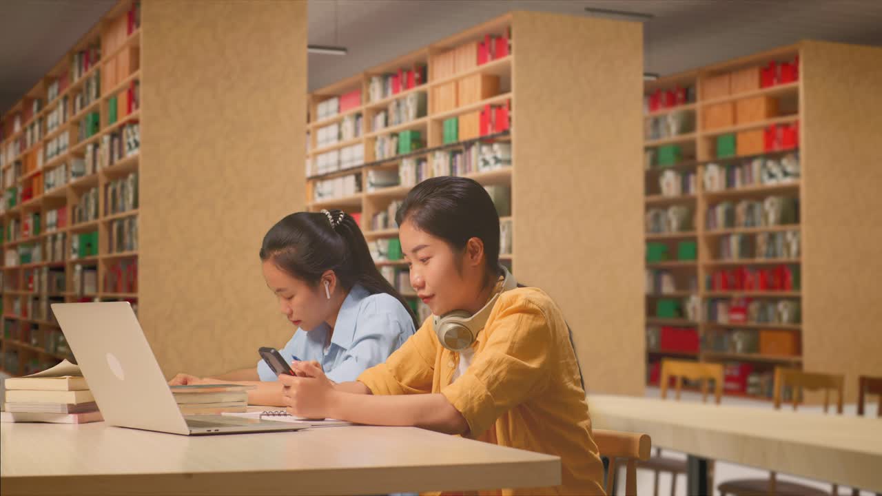 Asian Woman Student With Headphones Enjoys Using Smartphone While Sitting With Her Classmate Studying On A Table With Laptop In The Library