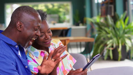 Smiling-Senior-Couple-Sitting-At-Home-Making-Video-Call-On-Digital-Tablet