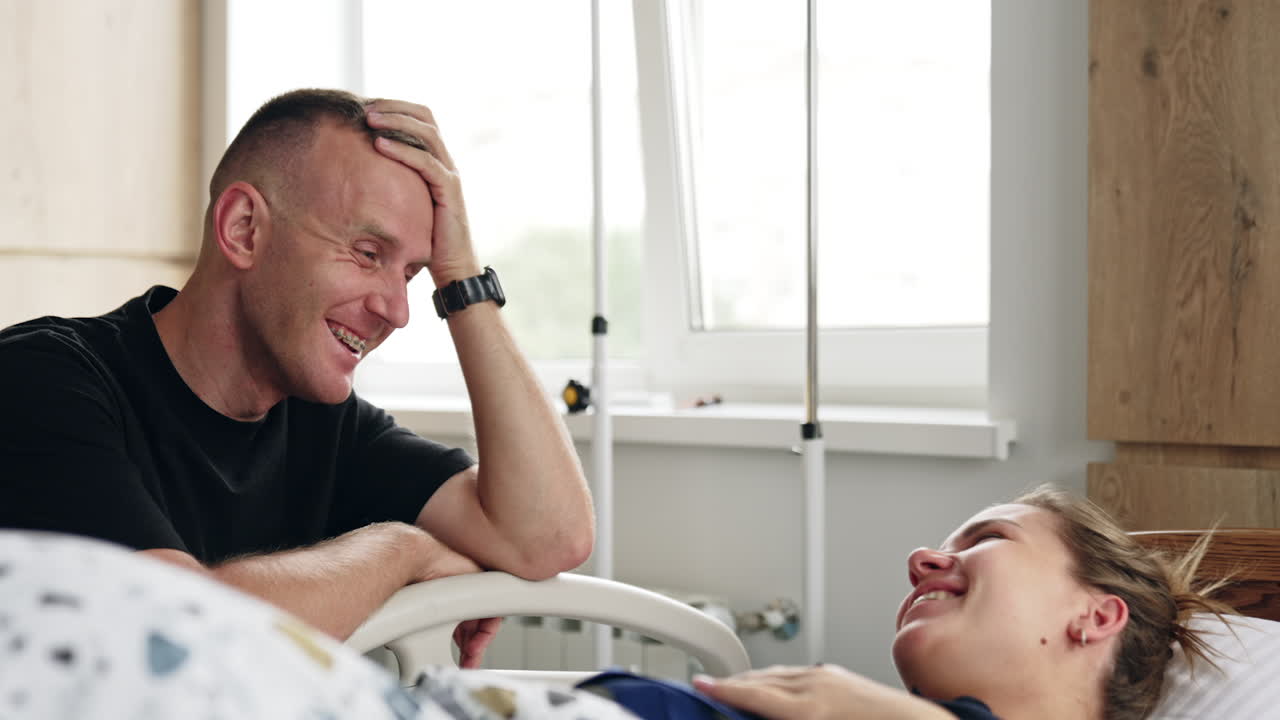 Man sits at the bed of a woman. Husband and pregnant wife chat and laugh while woman is checked with medical device.