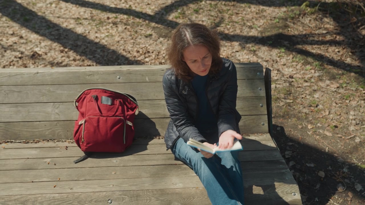 Woman reads book on wooden bench with red backpack beside her in park setting sunlight casts shadows on ground and subject. Woman wears dark jacket over blue shirt paired with jeans
