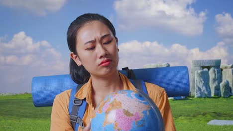 close up of asian female hiker with mountaineering backpack looking at world globe in her hands and making confused gesture while traveling in stonehenge