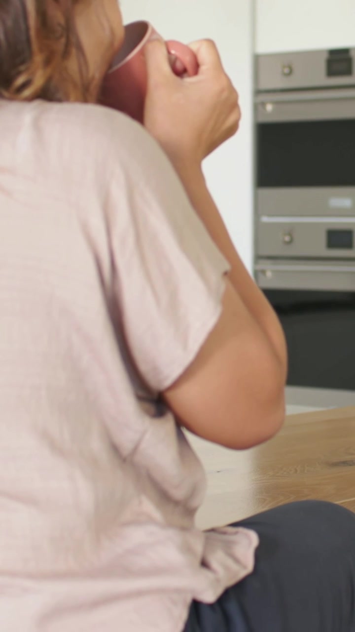 Video verticale: Una donna in maglietta rosa tiene in mano una tazza rosa, la porta alle labbra per sorseggiare, vicino al bancone della cucina e al forno.