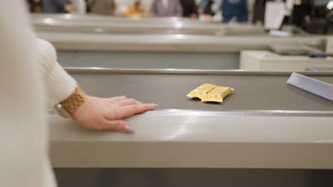 close up grocery shopper hand wearing wrist watch and polished nails resting on metal checkout counter edge with blurred conveyor belt and packaged energy bars in bright retail environment
