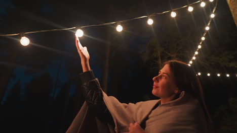 wife in blanket touches lights in glamping by forest. woman finds tranquility in hush and seclusion of camping experiencing blend of relaxation and solitude