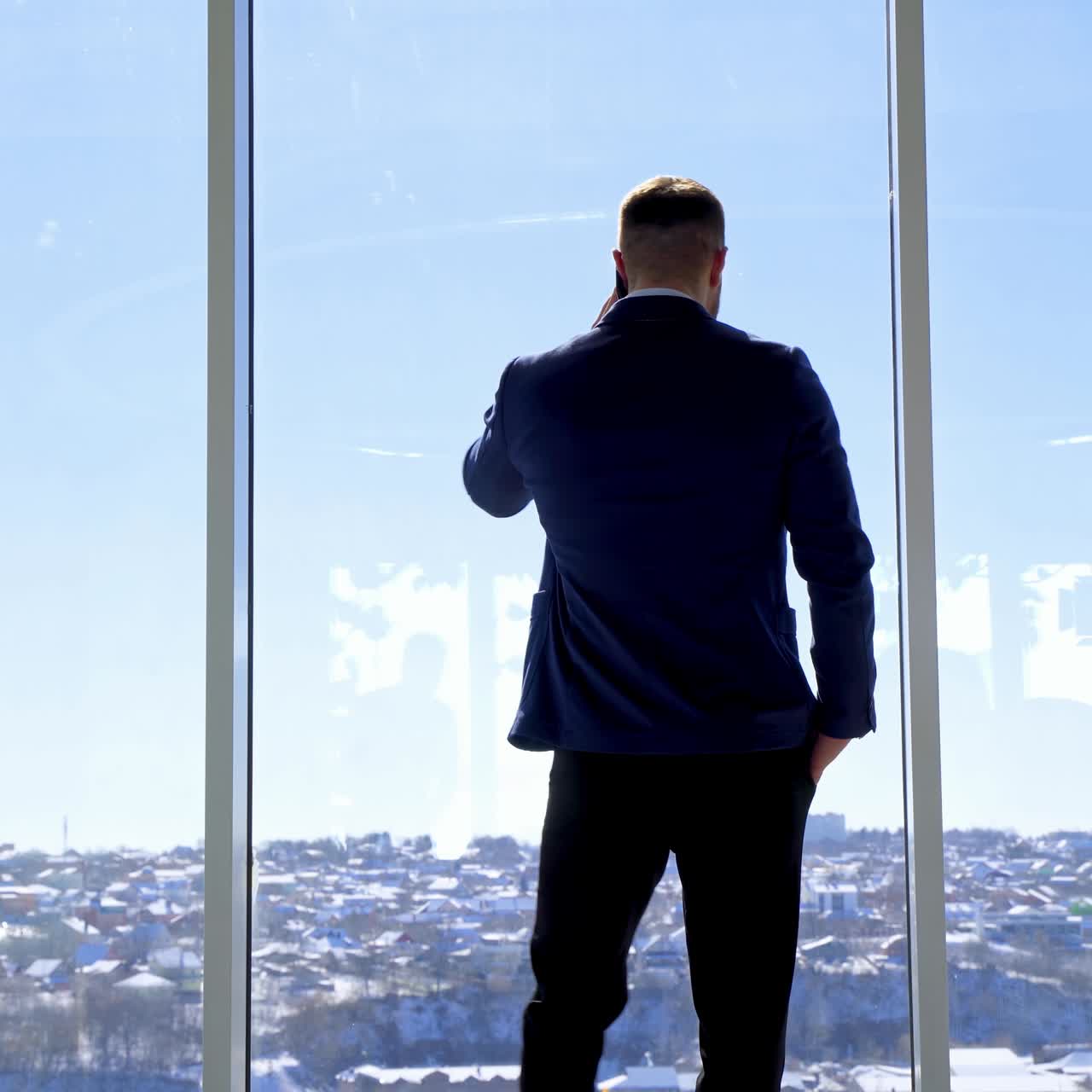 Businessman standing in his office. Rear view of a man wearing a suit talking the phone and looking out of the window. Panoramic window view with a city background