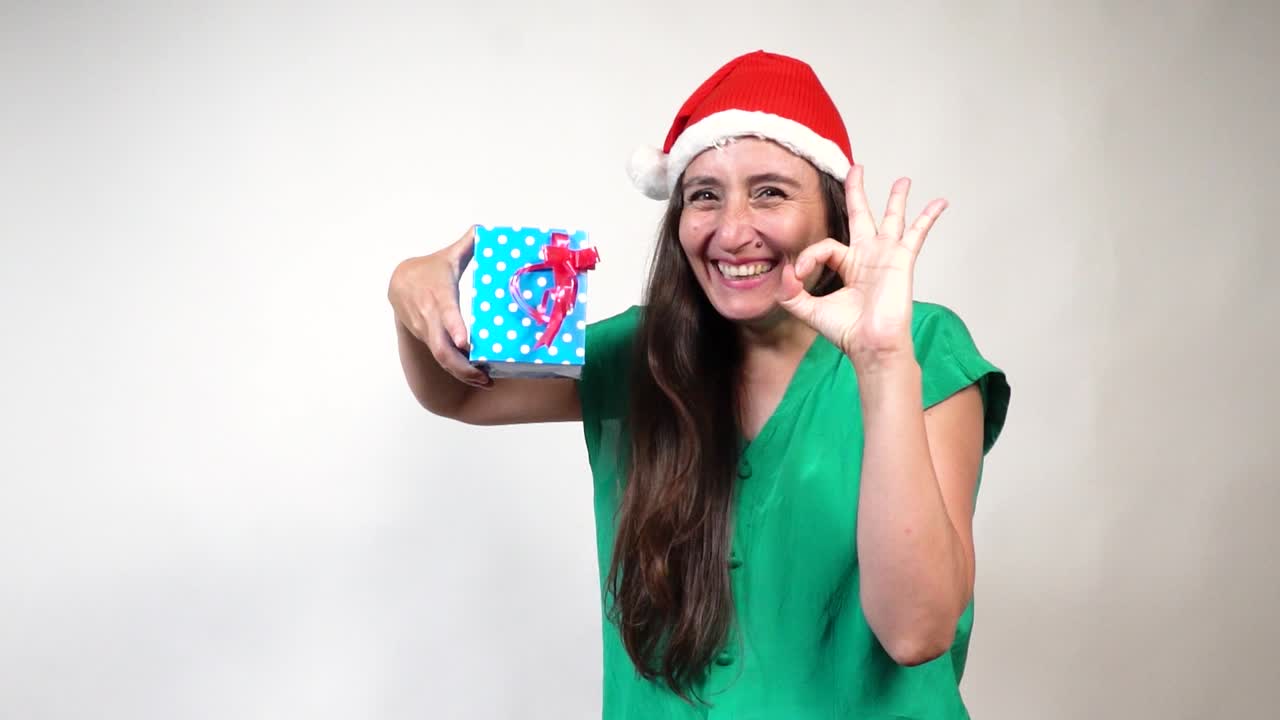 Cheerful brunette in Santa hat flashes OK gesture, holds blue polka-dot gift against white backdrop