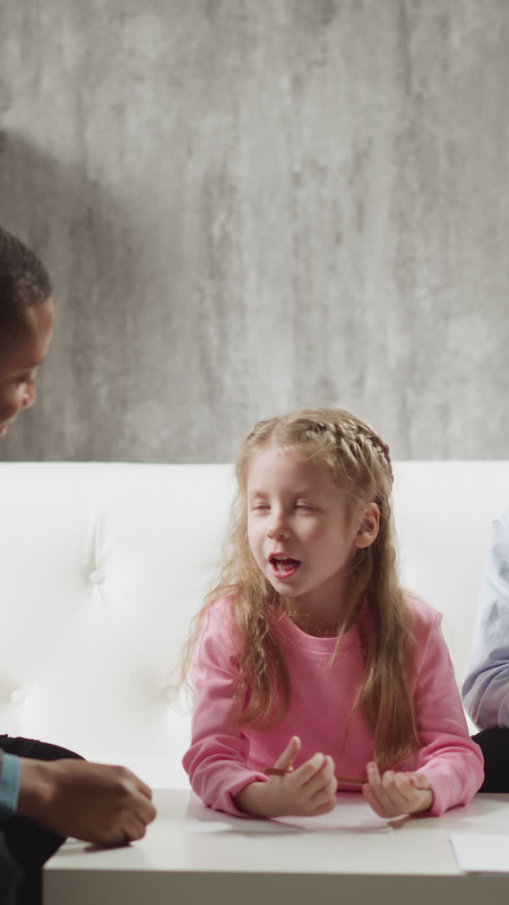 Cute little girl student tells new words to African-American teacher while brother sits nearby on sofa at home lesson with native speaker slow motion