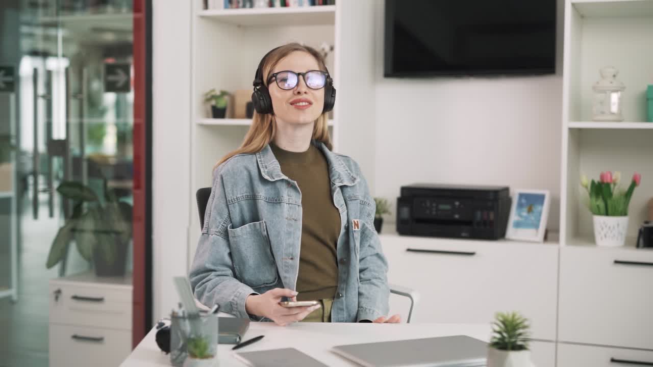 The woman chooses which song to listen to on her large wireless headphones. She sits at a table in a bright, modern room resembling an office