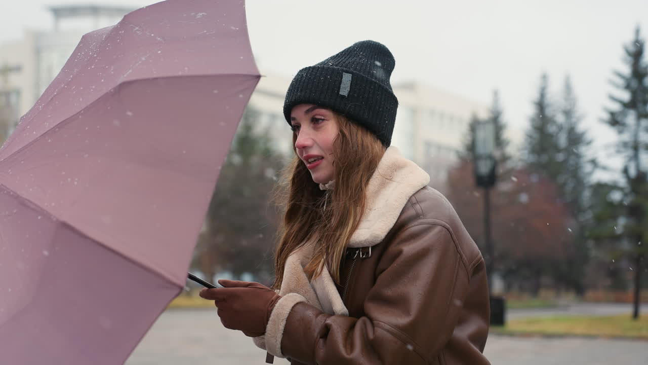Young girl holding umbrella wearing black knit cap and brown shearling jacket, surrounded by pine trees and urban buildings, with soft snowfall falling on cold overcast day