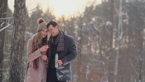 Happy-couple-holding-hot-tea-cups-over-winter-landscape.-Young-couple-in-love-on-a-winter-vacation,-standing-next-to-a-tree-and-drinking-hot-cup-of-tea