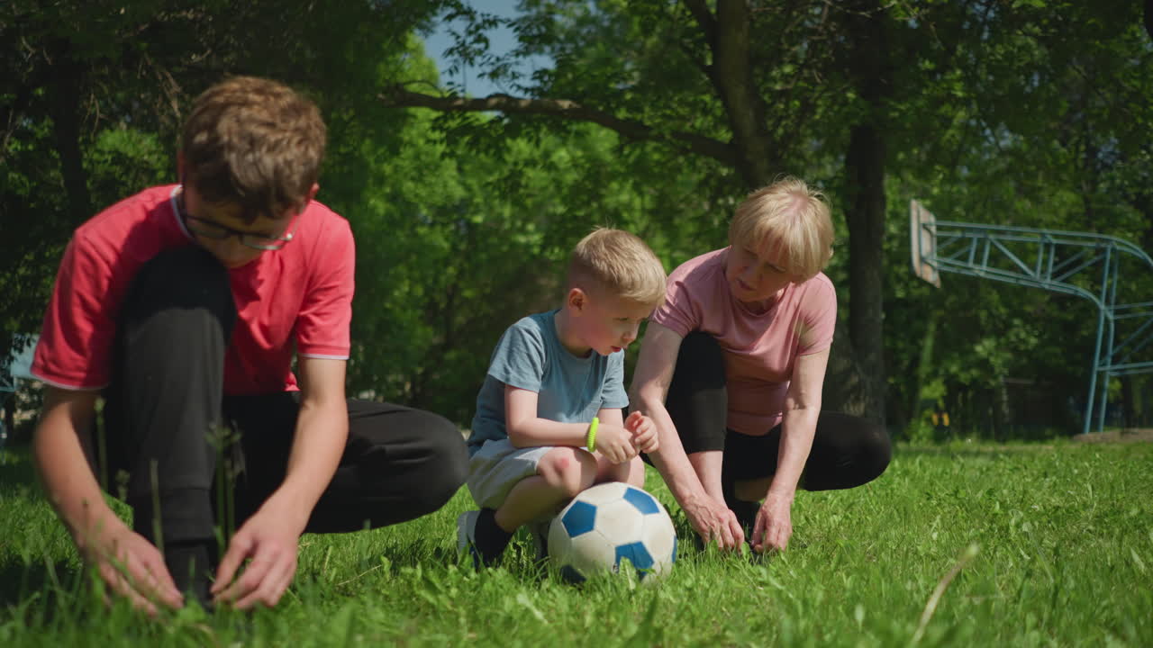 Close-up of a family moment outdoors where a mother and her older son tie their shoelaces on a grassy field while the younger boy balances on a soccer ball, looking towards his mother