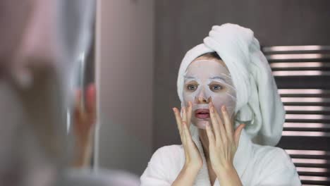 Woman-in-bathrobe-and-towel-with-white-mask-for-moisturizing-on-face,-looking-at-the-mirror