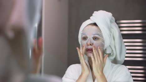 woman in bathrobe and towel with white mask for moisturizing on face, looking at the mirror