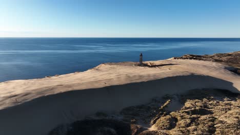 captured in denmark during a sunny evening, this wide angle drone shot reveals vast sand dunes and a historic stone lighthouse, showcasing the serene coastal landscape bathed in golden light.