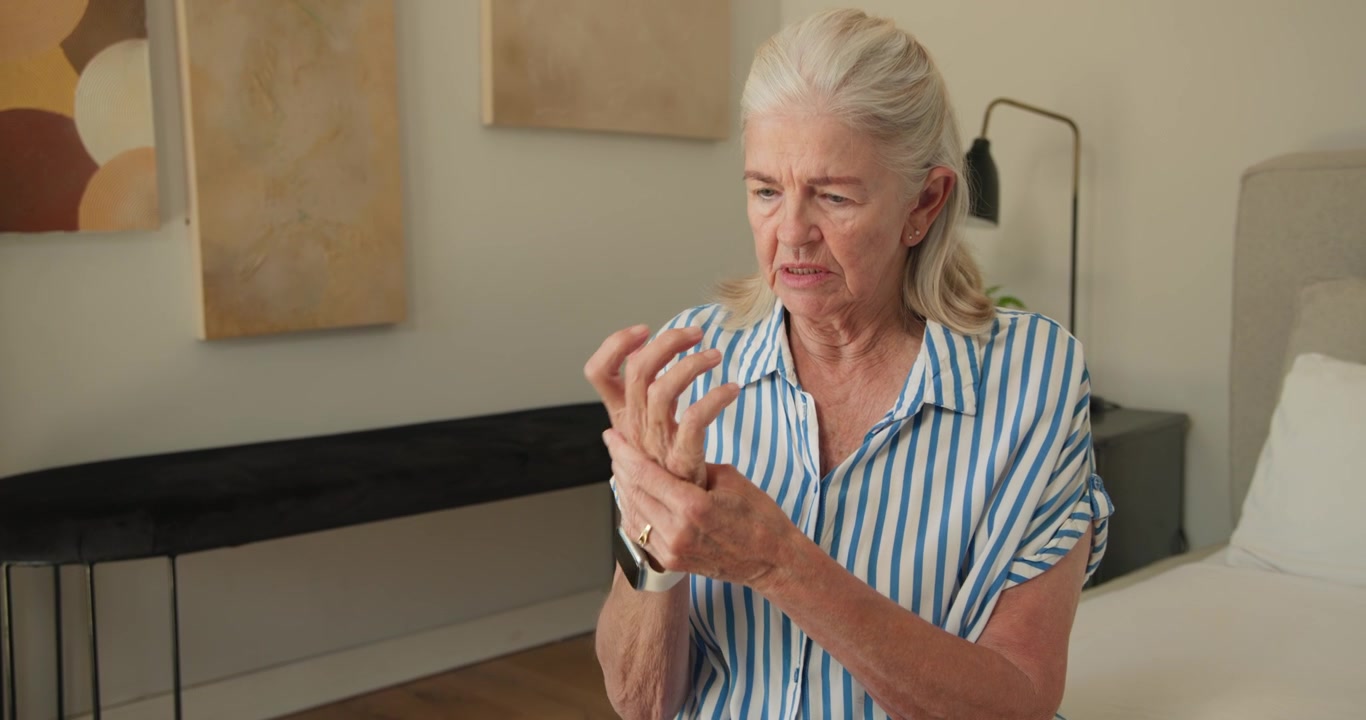 Anziana donna che si tiene il polso sinistro dopo aver sentito dolore, strofinando e controllando sotto lo smartwatch sul letto.