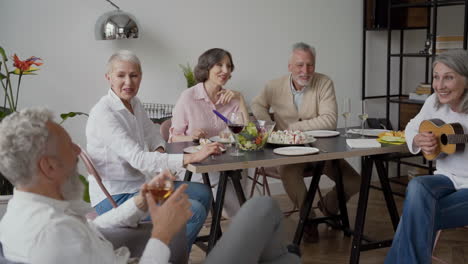 happy senior woman singing and playing the guitar sitting on chair, while her elderly friends listening to her and singing together sitting at the table